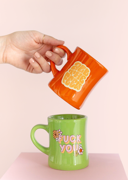 girl holding a tilted orange old fashioned diner-style mug with saying "all the feels" in lighter orange with yellow shadow over top of a green old fashion diner mug which says "fuck you" in pink with flowers
