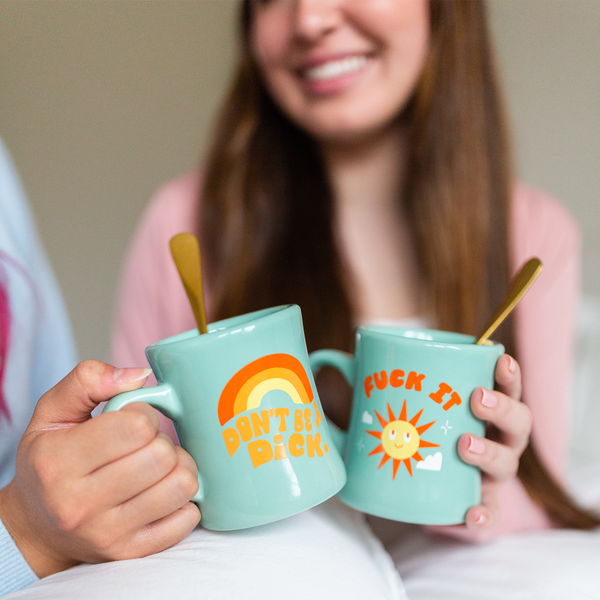 Two mint-colored mugs. 1 has the phrase, "Don't be a Dick" printed on with a rainbow arch printed over the phrase and the other mug has the phrase, "Fuck It," printed on with a smiley sun and clouds printed beneath the phrase. Both mugs are being held up by two people in the background.