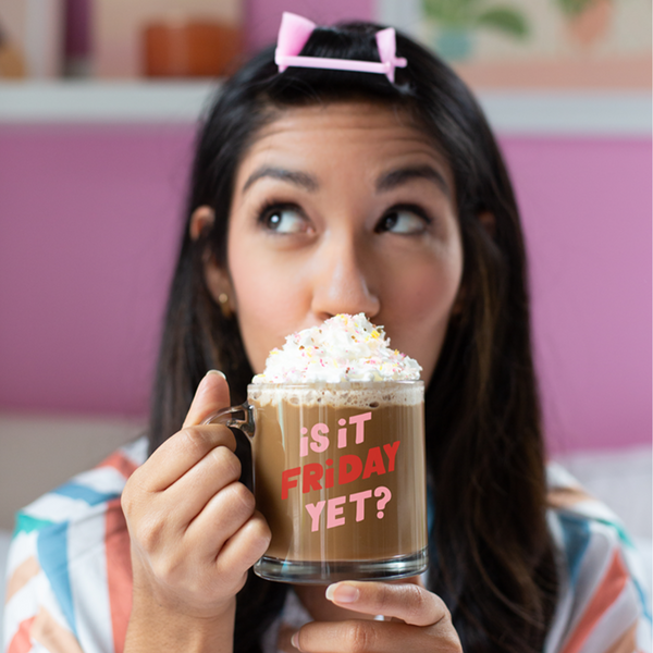 Brunette woman with a pink hair roller, holding a is it friday yet clear glass mug with pink and red lettering sipping on hot chocolate with whipped cream and sprinkles.