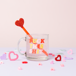 cute party scene with fuck this shit glass mug filled with a carbonated drink and has a glitter heart drink stirrer laying diagonal in the glass with pin purple and red hearts on the purple table with a pink background.