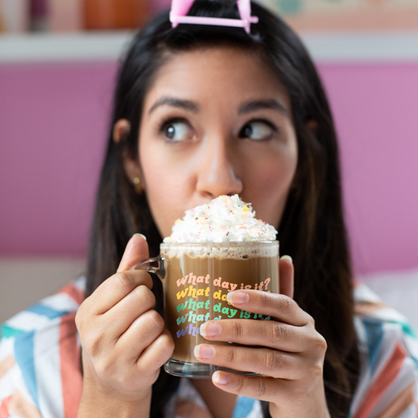 A person holding a "What day is it?" glass mug with hot chocolate and whipped cream in it. Wording on mug is repeating 5 times with different colored lettering in each repitition.