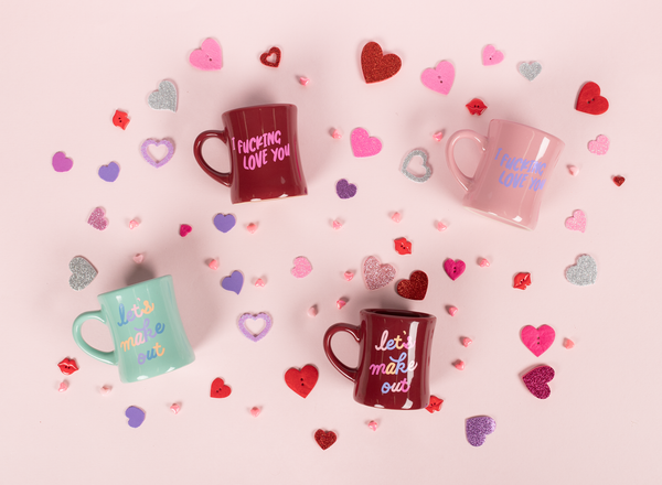 Diner mugs in sangria, mint, and blush. The mint mug has the words "let's make out" in alternating colors. Sangria mugs have "I fucking love you" and "let's make out." Blush mug has "I fucking love you" in periwinkle writing. The mugs are surrounded by felt craft hearts and heart beads in purple, pink, silver, and red.