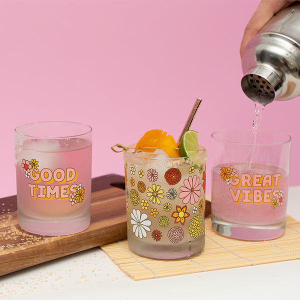 person pouring water into a set of 4 retro flower power rocks clear glass set sitting on a white table with a pink wall in the background.
