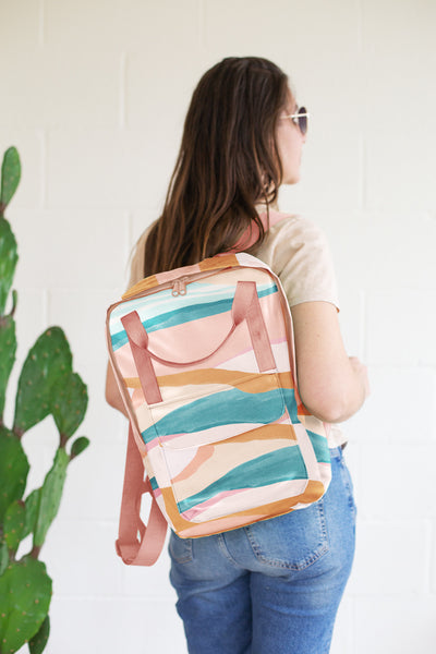 Vegan leather backpack with peach webbing and landscape pattern. Displayed by person holding back pack over their shoulder in front of white background.