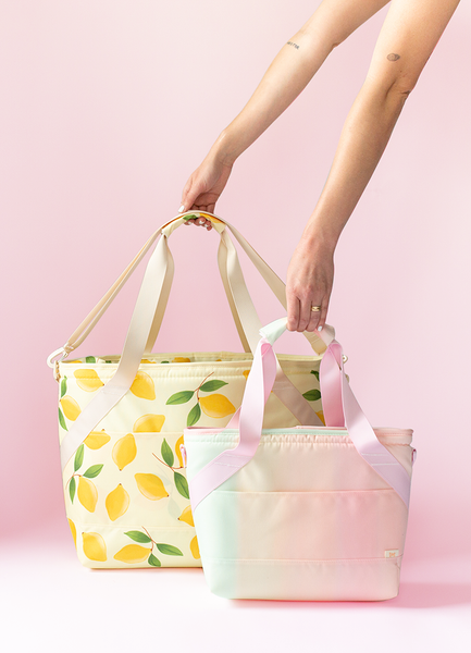 Lady holding soft yellow cooler bag with a print of lemons next to a smaller gradient pastel cooler bag. 