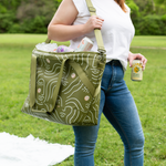 woman in blue jeans with white shirt carrying Ice Queen Zen Ladies shoulder cooler tote with olive green background and white retro ladies stencil drawing with pink and white cheeks.