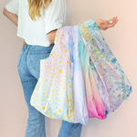 A person holding 4 differently designed Twist and Shout tote bags. One bag is designed with white daisies, next is a light blue/purple tie dye. Next is a rainbow ombre colore, and the last is a multicolored floral bag withs blues, purples, and oranges.