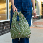 An olive green  tote bag with female faces printed on in white lines that are not connected being held by a person in front of a store front.