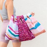 Three tote bags (Twist and Shouts) being held up by a person. The left bag is an abstract, multicolored line design. The middle is a Terrazzo-speckled design with a magenta background color. The right is an overall wave-like design with pastel and neutral colors.