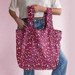 A multicolored, jewel toned Terrazzo speckled tote bag with a magenta background. Bag is being held by a person in front of a pink background.