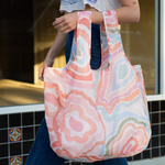 A person wearing a large Sguiggles Twist and Shout which is a multicolored tote bag with a water stain design. Colors are pastel oranges, neutral blues and reds, along with neutral greens, creams, and browns.