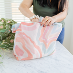 A person using a Squiggles Twist and Shout which is a multicolored tote bag with a water stain design. Colors are pastel oranges, neutral blues and reds, along with neutral greens, creams, and browns.