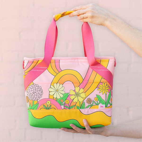 A cute, groovy flower pattern with rainbow swirls printed onto the tote-like shaped bag being held up by a pair of hands in front of a light background.