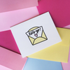 White greeting card with a yellow, black and pink graphic. The graphic is a yellow envelope with a paper sticking out with script text 