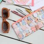 A vinyl clear pixie pouch with pastel multicolored moon phases printed on. Also has three Jotter pens in the colors Gold, Rose Gold, and Silver. Displayed with notebooks and sunglasses.
