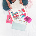 Girl writing in a journal surrounded by books, magazines, and a powder blue splatter laptop sleeve.