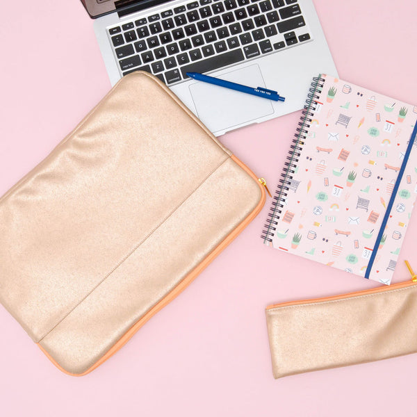 A gold-metallic colored laptop sleeve with a slip pocket on the side. Displayed with a laptop, a Jotter Pen, a Lucky charms notebook, and a metallic-gold pixie pouch, all on top of a muted pink background.