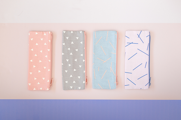 Four pouches sit on a surface. One with peach canvas material and white triangles. The second a grey background with white triangles. The third is a washed out denim with pink marks, and the fourth pouch is a light pink with navy marks.  