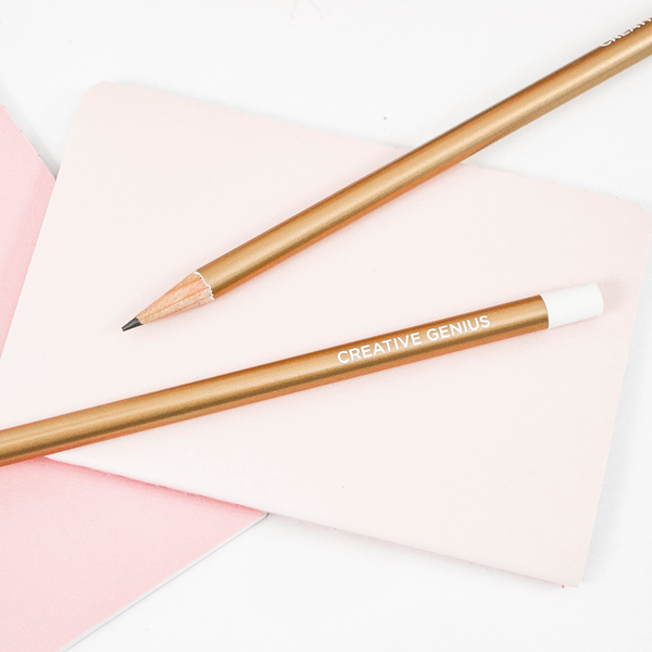 Two gold pencils with the text "Creative Genius", laying on a white and pink surface