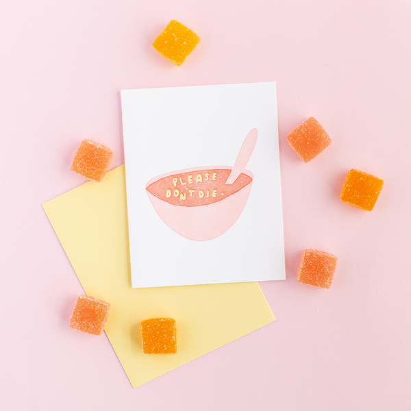 White greeting card with a pink bowl of letter soup. The letters are arranged into "Please Don't Die". There is a yellow envelope, orange square candy and a pink background.