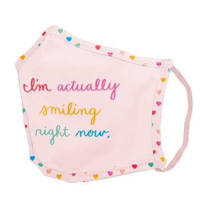 A pink facemask with rainbow themed tiny hearts on the edges, with "I'm actually smiling right now" written in color alternating scripts. 