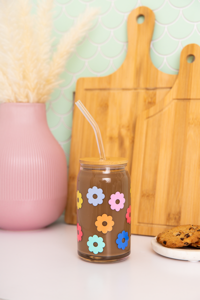 Can glass with straw and lid with multi-color daisies wrapping the glass; glass is filled with liquid and surrounded by various items.