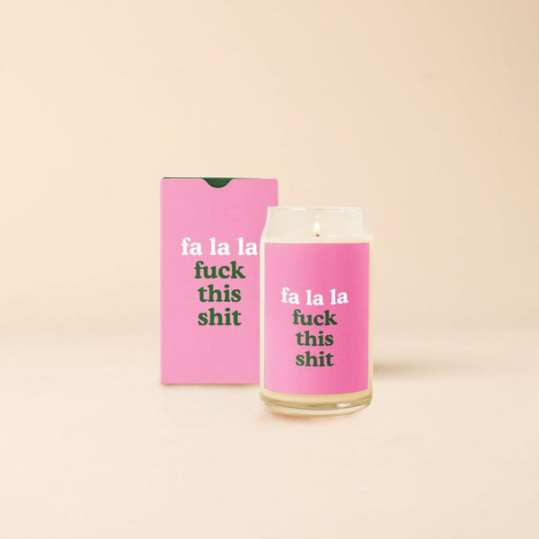 Holiday candle with pink decal that says,"fa la la fuck this shit." Displayed with the packaging box that is pink with the same phrase printed in the front.