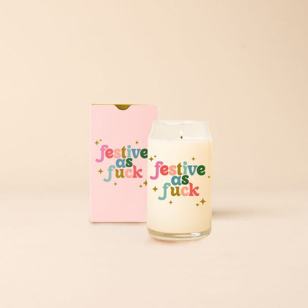 A 12 oz. candle that says "Festive as fuck," on the front with multicolored lettering. Candle is displayed with packaging box that is a light pink and also has "Festive as Fuck" printed on the front in the same lettering.