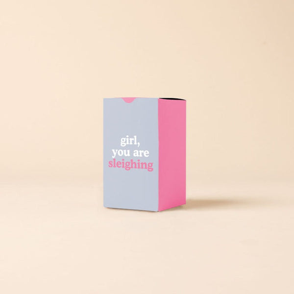 A candle box that is pink on three sides and periwinkle on the front side. Front side says, "girl, you are sleighing."