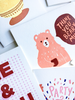 An assortment of white greeting cards partially cropped. The main card has a peach colored bear holding a red mug. There is a text bubble with 
