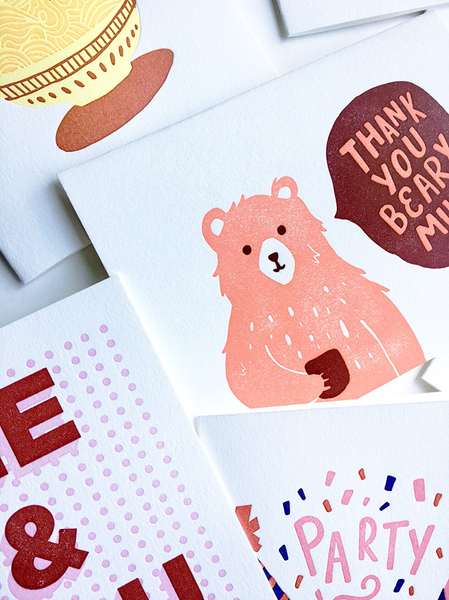 An assortment of white greeting cards partially cropped. The main card has a peach colored bear holding a red mug. There is a text bubble with "Thank You Beary Much". 