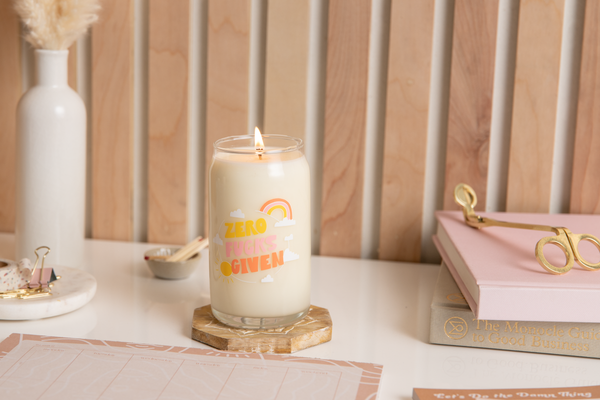 Candle can glass with text that reads "ZERO FUCKS GIVEN" in yellow, pink, and orange font. Text is surrounded by illustration of a small rainbow and clouds. Candle is surrounded by various stationery.