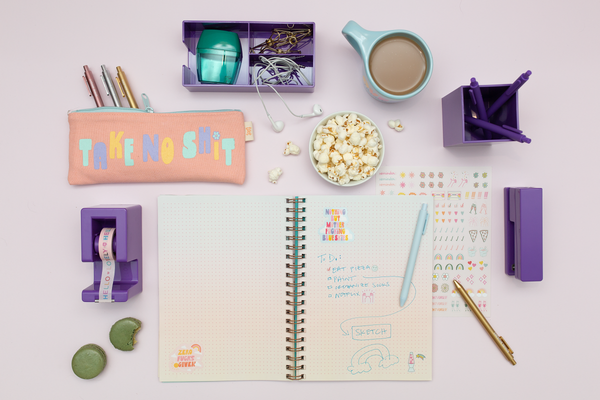 Bullet Journal with powder blue jotter pen writing. Also pictured is a gold jotter pen, purple desk set items, Over Thinker element mug and Take No Shit pixie pouch. 