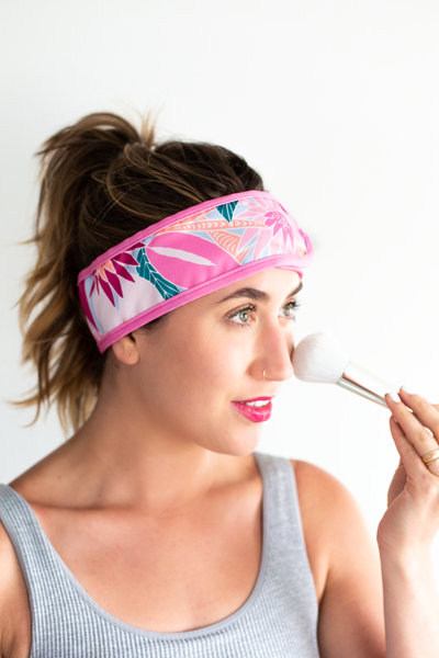 A woman wearing the Floral Day spa head wrap, and holding a makeup brush to her cheek. The spa head wrap has a pink trim and a floral pattern.