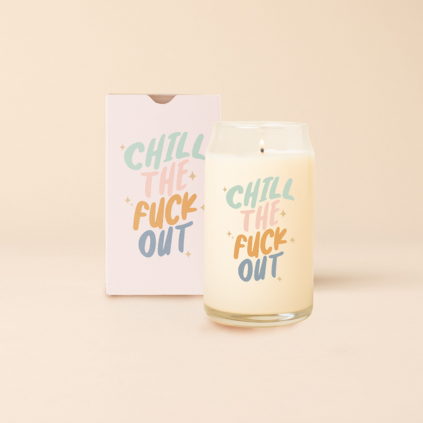 Can glass candle with text that reads "CHILL THE FUCK OUT" in multi color font, with minimalist sparkle stars surrounding text. Box packaging with the same design sits behind candle.