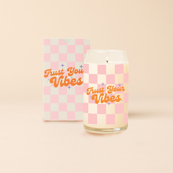 Can glass candle with pink checkered print wraps around the candle, with text that reads "TRUST YOUR VIBES" in orange font, text is surrounded by purple minimalist sparkle stars. Box packaging with same design sits behind candle.