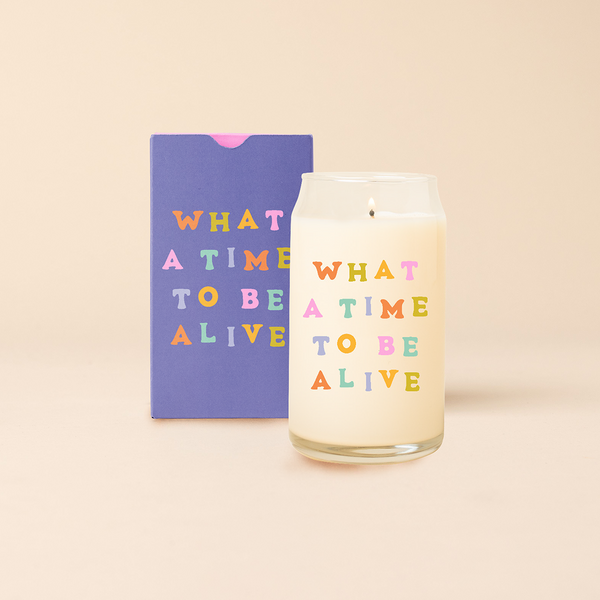 Can glass candle with text that reads "WHAT A TIME TO BE ALIVE" in multi-color font. Box packaging with same design sits behind the candle.