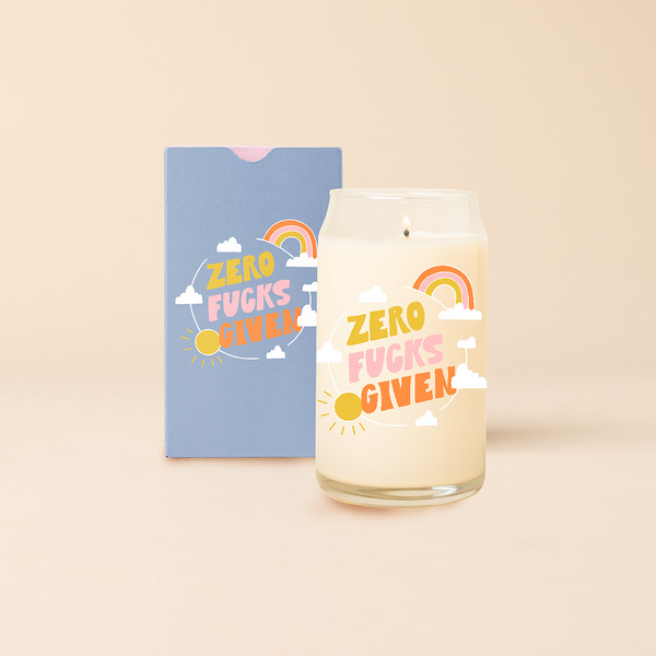 Can glass candle with text that reads "ZERO FUCKS GIVEN" in yellow, pink, and orange font. Text is surrounded with a small rainbow, clouds, and illustration of the sun. Box packaging with same design sits behind candle.