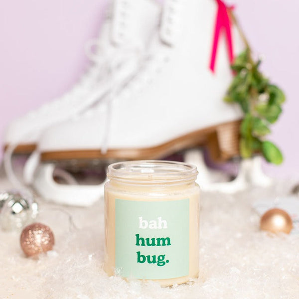 A Holiday Candle Jar with a mint green decal that says "bah hum bug," in white and dark green colored text. Displayed in front of ice skates.