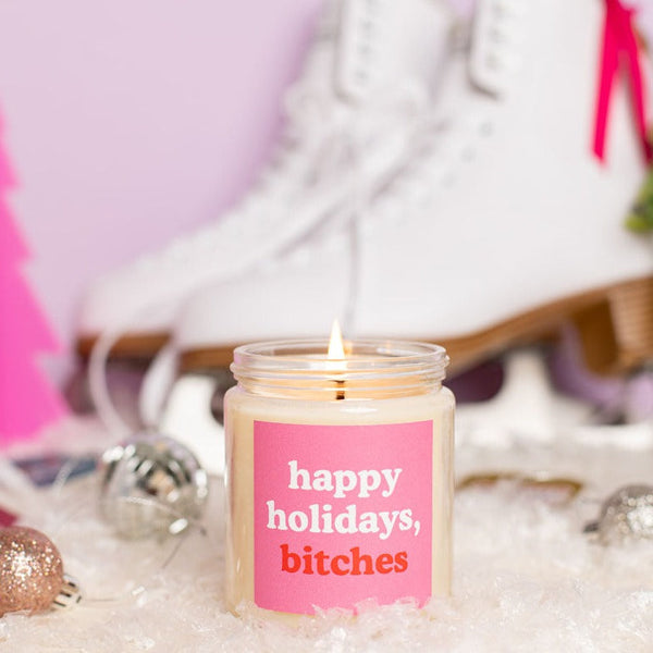 A TOOT Holiday Candle Jar with a pink decal that says," happy holidays, bitches." Candle is displayed in front of skates.