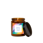 Amber glass candle jar with sticker on front with multi-colored scallop design and text that reads "HAPPY YOU'RE HERE" in blue font in a white square.