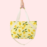 A large tote bag with a lemon and lemon leaf design. Bag has side pocket and white straps. Tote bag is being displayed in front of a white background.