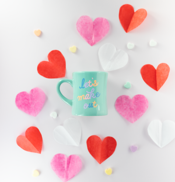 Mint diner mug with the cursive words "let's make out" in alternating colors. The diner mug is surrounded by paper hearts in pink, red, and white, as well as candy conversation hearts.