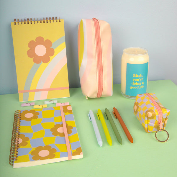 Light blue background and a rainbow flower pattern with green taskpad, rainbow wave jitterbug, "bitch you're doing a good job" candle, cool funky daisy notebook, jotter pens and a cool funky key ring on a pastel color surface.