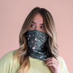 A multicolored terrazzo speckled mask-like item with a dark teal background color. Displayed on a person wearing it from their nose to their neck in front of a muted pink background.