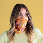 An orange mask-like item with multicolored and differently designed flowers. It covers from the nose to the bottom of the neck and is displayed by a person wearing it in front of a yellow background.