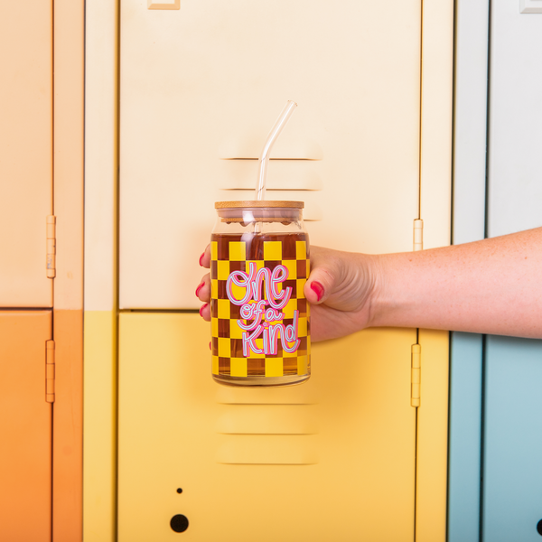 16 oz can glass with lid + straw. Yellow checker print wraps around the glass, text that reads "One of a Kind" is on the front of the glass in blue and pink font. A hand is holding the glass in front of lockers.