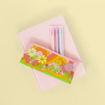 Cute pencil pouch in pink with flowers and swirls  and a pink zipper on top of a light pink notebook and a set of three jotter pens in light pink, lilac and baby blue coming out of pouch.