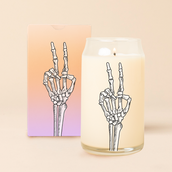A 16 oz candle can glass with an illustration of skeleton hand holding up a peace sign. An orange-to-purple ombre box with the same illustration sits behind the candle.