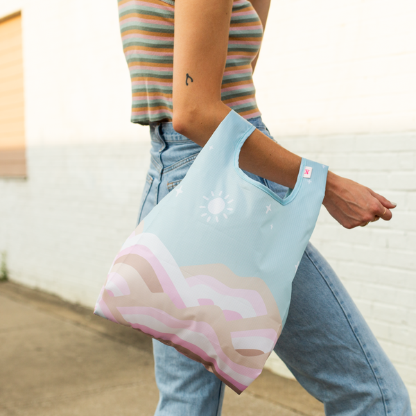 Woman carrying a small sized pastel blue reusable tote bag with pink, brown, and white hill designs on the front and back of the bag.